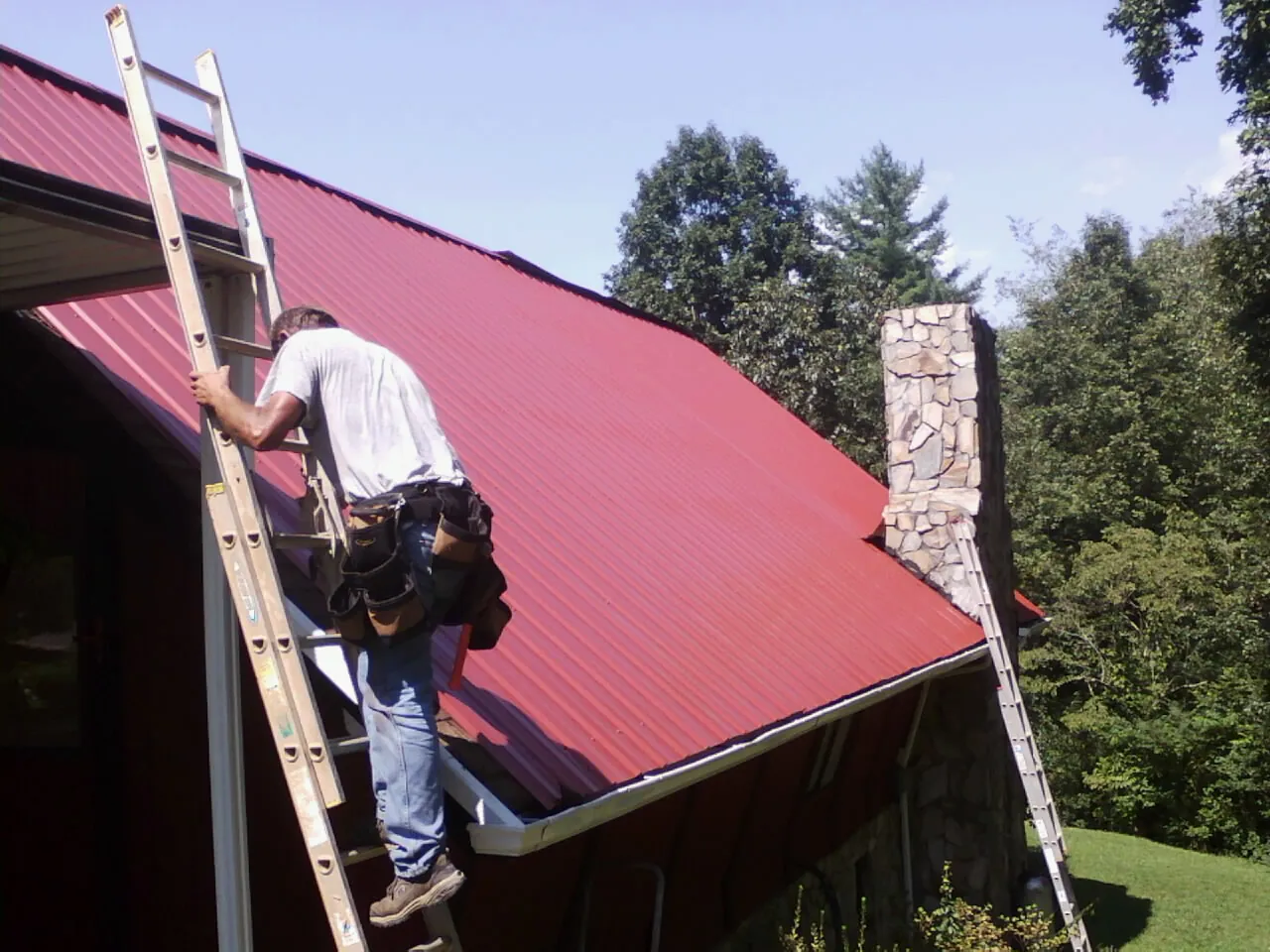 Roofer on a ladder beside a red metal roof and stone chimney — Soffit & Fascia Repair by Roofing Brothers in Meredith